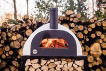 Load image into Gallery viewer, A wood fired pizza oven lit and placed in front of a stack of timber
