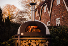 Load image into Gallery viewer, A pizza oven on top of a pizza oven stand that is lit, placed in front of a brick house
